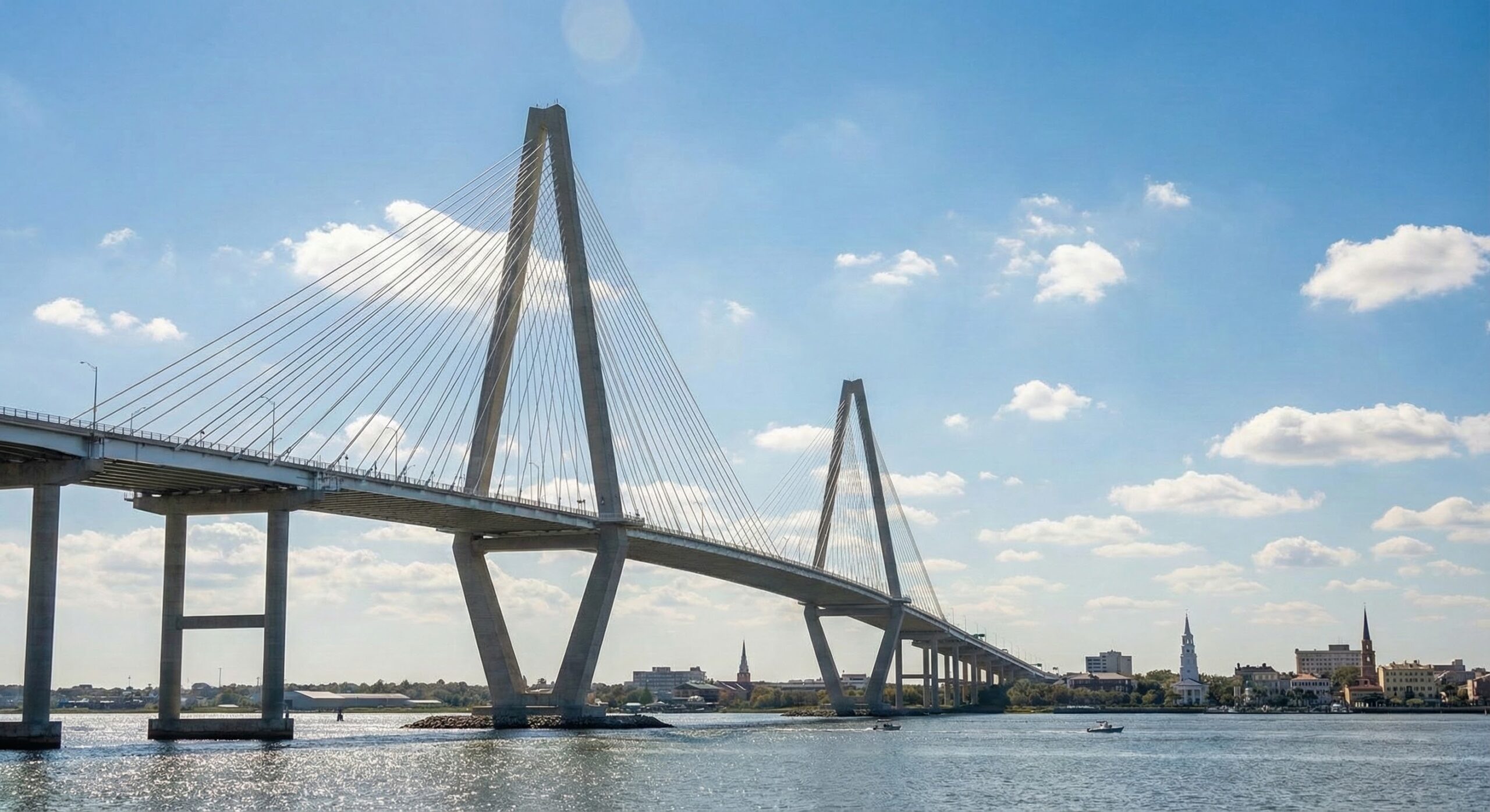 A large cable-stayed bridge spans across a wide river under a blue sky with scattered clouds, with small boats on the water and a city skyline visible in the distance.