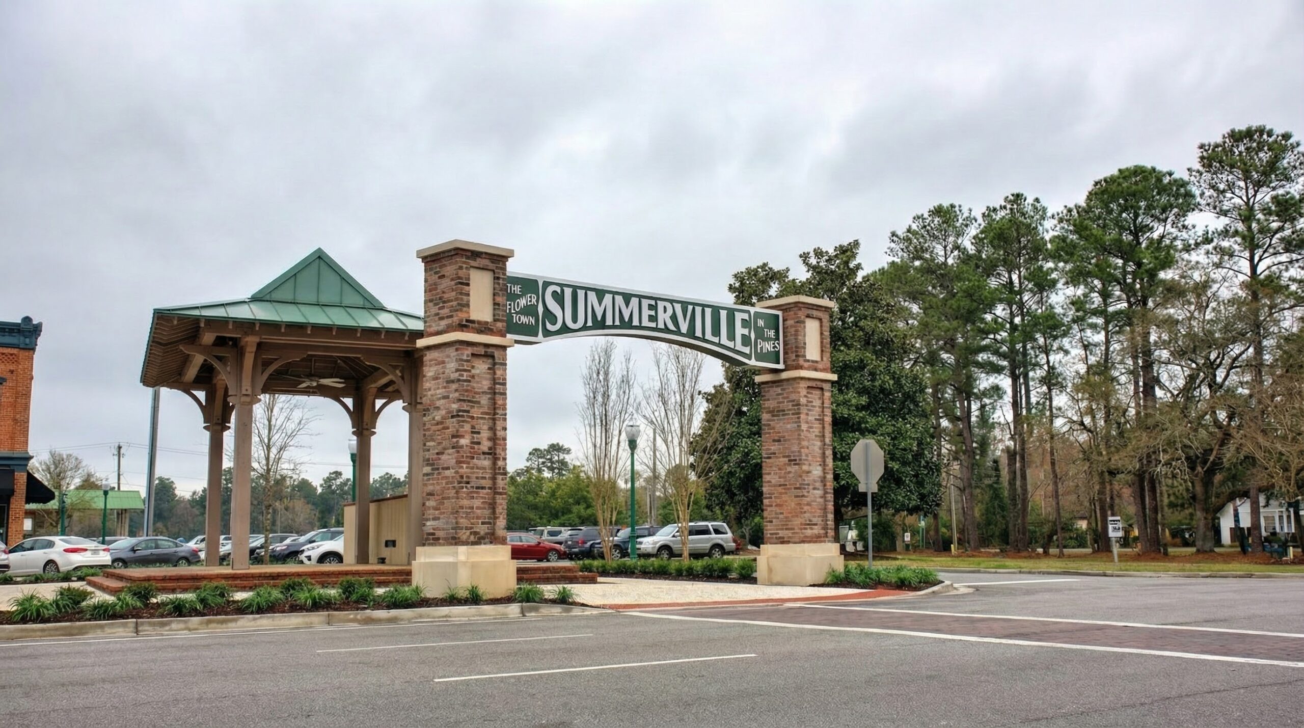 A large brick and wood entrance sign reads Summerville over a road, with parked cars, trees, and buildings visible in the background under a cloudy sky.
