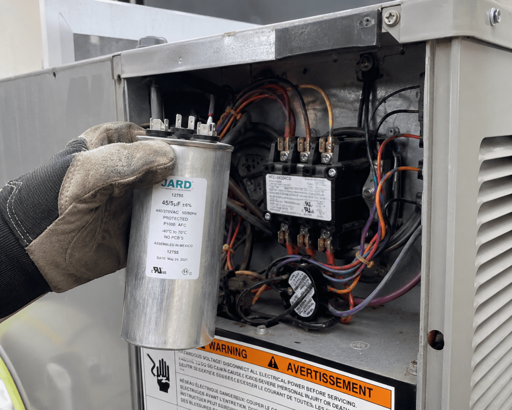 A gloved hand holds a metal capacitor near exposed wiring and components inside an HVAC unit’s electrical panel. Warning labels are visible, highlighting potential AC capacitor failure symptoms and causes during inspection.